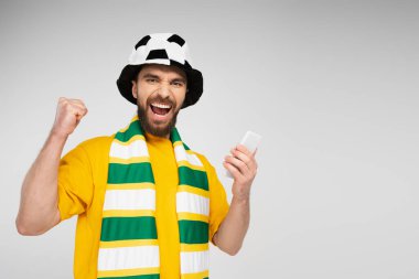 Futbol şapkası ve çizgili atkısı olan neşeli bir adam akıllı telefon tutuyor ve gri renkte mutlu bir şekilde izole edilmiş.