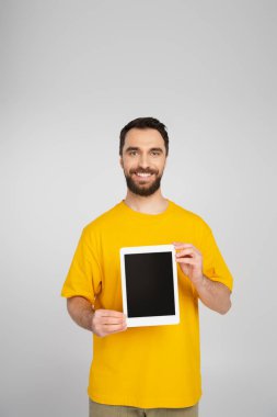 Kumral sakallı adam, dijital tableti boş ekranla gösteriyor ve gri ekranda izole edilmiş kameraya gülümsüyor.