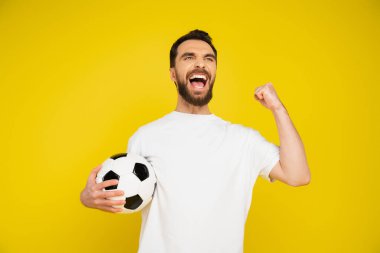 overjoyed football fan with soccer ball screaming and showing win gesture isolated on yellow clipart