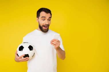 Şaşkın ve mutlu bir spor fanatiği futbol topuyla cep telefonundan izlenen futbol maçını sarı renkte izliyor.