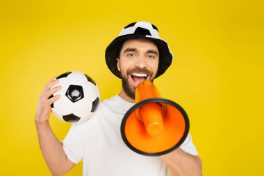 Futbol fanatiği şapkalı neşeli adam sarıda izole edilmiş megafonla bağırıyor.