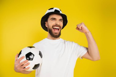 Futbol topunu tutan şapkalı haykıran futbol fanatiği ve sarıda izole edilmiş başarı jesti