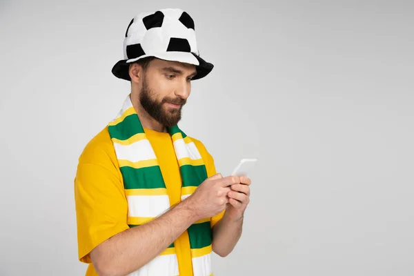 focused man in football fan hat and striped scarf making sports betting on smartphone isolated on grey