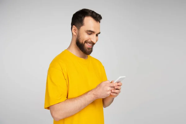 グレーに隔離された携帯電話を使って黄色のTシャツを着た髭の男を笑っています — ストック写真