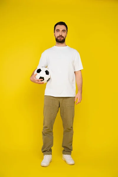 全身上下都是黑发 留着胡子 身穿白色T恤和米色裤子的男人 站在黄底足球旁边 — 图库照片