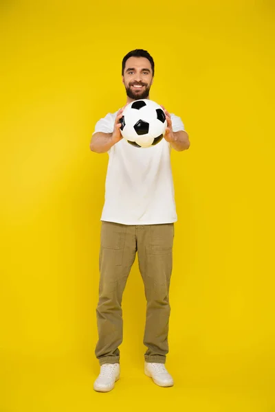 full length of pleased man in pants holding soccer ball in outstretched hands on yellow background