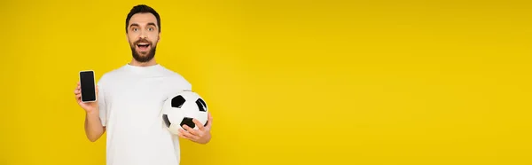 サッカーボールとスマートフォンを持つ驚きのスポーツファン黄色 バナーに隔離された空白の画面で — ストック写真