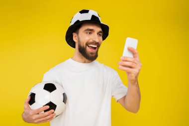 Şapkalı spor taraftarı, sarı telefondan izole edilmiş akıllı telefondan futbol maçı izliyor.