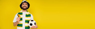 happy man in football fan hat standing with credit card and soccer ball isolated on yellow, banner clipart