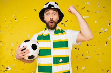 amazed football fan in hat and scarf showing triumph gesture under falling confetti on yellow background clipart