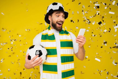 Futbol topuyla hayretler içinde ve mutlu bir sporcu futbol maçı izliyor. Akıllı telefondan, sarı konfeti yakınlarında. 