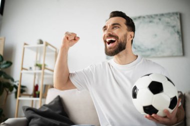 shouting bearded man holding soccer ball and showing win gesture while watching football match at home clipart