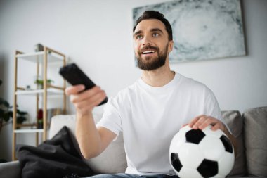 Mutlu adam evde şampiyonayı izlerken elinde futbol topu ve uzaktan kumanda tutuyor.
