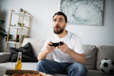 KYIV, UKRAINE - 21 Ekim 2022: evde pizza ve bira şişesinin yanında video oyunu oynayan gergin adam