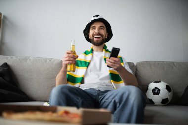 Fan şapkalı ve fularlı mutlu bir adam futbol maçı izlerken elinde uzaktan kumanda ve bir şişe bira tutuyor. 
