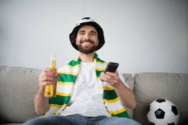 Hayran şapkalı ve atkılı neşeli bir adam futbol maçı izlerken elinde uzaktan kumanda ve bir şişe bira tutuyor. 