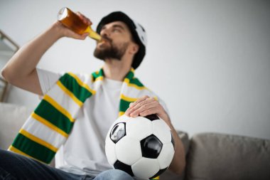 Şapkalı ve atkılı sakallı adam bira içerken elinde futbol topu tutuyor. 