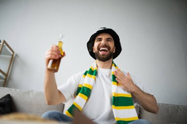 Atkısı ve şapkası olan hayret verici sporsever fanatiğin düşük açılı görüntüsü şampiyonluğu izlerken elinde bir şişe bira tutuyor.
