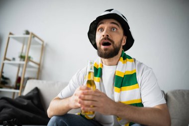 KYIV, UKRAINE - OCTOBER 21, 2022: astonished man in scarf and hat holding bottle of beer while watching championship clipart