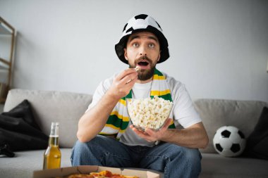 KYIV, UKRAINE - 21 Ekim 2022: Sportif fan şapkalı ve atkılı şok adam patlamış mısır yiyor ve şampiyonayı seyrediyor. 