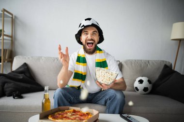 KYIV, UKRAINE - 21 Ekim 2022: Fan şapkalı heyecanlı adam lezzetli yemek ve bira şişesinin yanında şampiyonluk maçı izliyor.