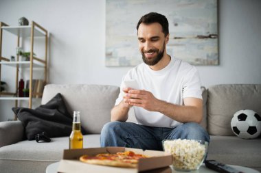 KYIV, UKRAINE - 21 Ekim 2022: Kutuda bira ve pizza şişesinin yanında akıllı telefon kullanan neşeli adam 