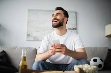 cheerful man using smartphone near bottle of beer and tasty food on blurred foreground  clipart