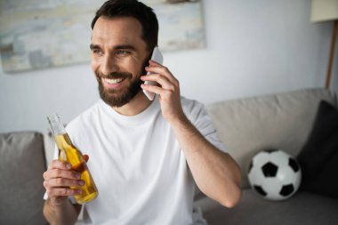 Neşeli bir adam elinde bira şişesiyle futbol masasının yanındaki bulanık koltukta akıllı telefondan konuşuyor. 