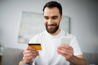 Evde online alışveriş yaparken kredi kartı tutan ve akıllı telefon kullanan neşeli bir adam.