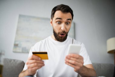 Evde online alışveriş yaparken elinde kredi kartı tutan ve akıllı telefon kullanan şok bir adam.