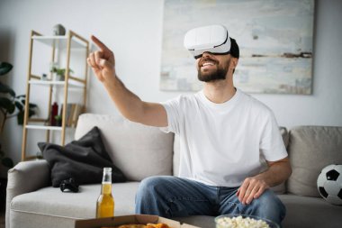 KYIV, UKRAINE - 21 Ekim 2022: VR kulaklıklı neşeli adam evde oyun oynarken parmağıyla işaret ediyor