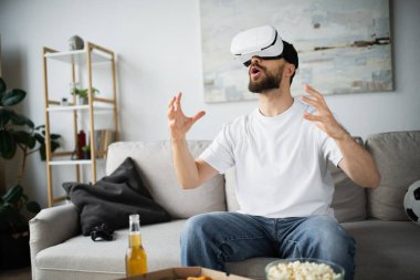 KYIV, UKRAINE - 21 Ekim 2022: VR kulaklıklı sersemlemiş sakallı adam evde oynarken el kol hareketi yapıyor