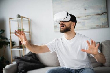 KYIV, UKRAINE - 21 Ekim 2022: VR kulaklıklı hayret ve sakallı adam oturma odasında oynarken el kol hareketi yapıyor