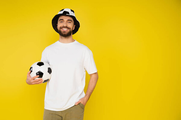 веселый футбольный болельщик в шляпе держа мяч и стоя с рукой в кармане изолированы на желтый