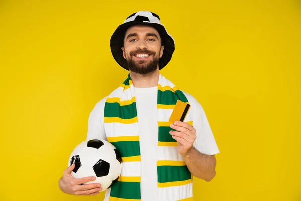 pleased football fan in hat and scarf holding ball and credit card isolated on yellow
