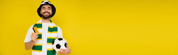 happy man in football fan hat standing with credit card and soccer ball isolated on yellow, banner