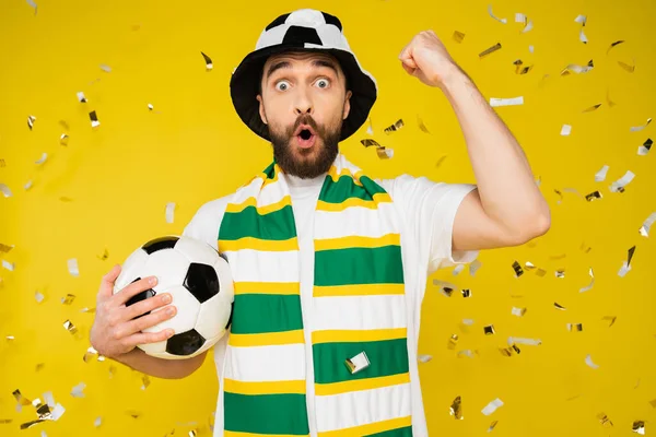 amazed football fan in hat and scarf showing triumph gesture under falling confetti on yellow background