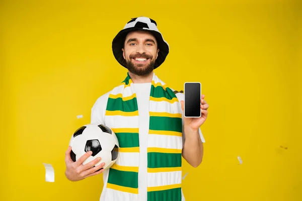 cheerful sports fan with soccer ball showing smartphone with blank screen on yellow background