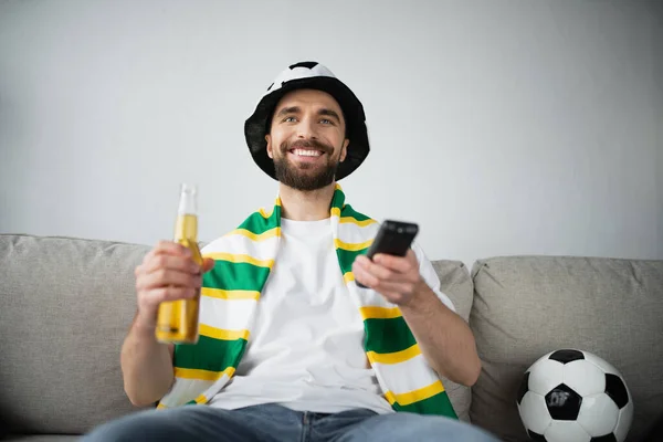 cheerful man in fan hat and scarf holding remote controller and bottle of beer while watching football game