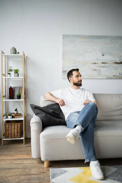 Vousatý Muž Džínách Sedí Gauči Moderním Obývacím Pokoji — Stock fotografie