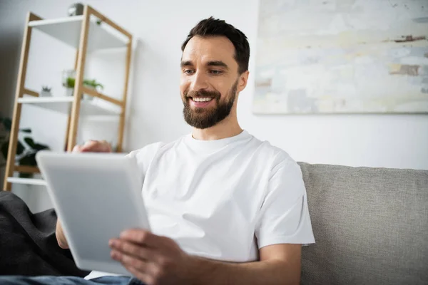 在现代客厅里 身穿白色T恤的快乐且满脸胡须的男人坐在沙发上 用数码平板电脑 — 图库照片