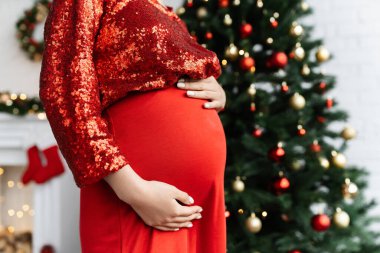 partial view of pregnant woman in elegant clothes touching belly near blurred Christmas tree clipart