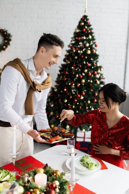 smiling man holding plate with grilled vegetables near asian wife with fork during romantic Christmas supper clipart