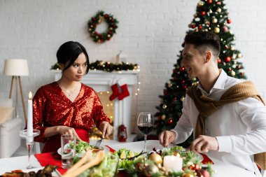 Mutlu adam, bulanık Noel ağacının yakınındaki şenlik yemeğinde hamile ve ciddi Asyalı eşine bakıyor.