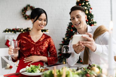 hamile Asyalı kadın bir bardak suyla kocasının yanında gülümsüyor Noel yemeği sırasında cep telefonunu gösteriyor