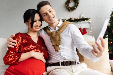 Hamile ve zarif Asyalı bir kadın. Kocasının gülümsemesiyle evde elinde Noel dekoru olan bulanık karta bakıyor.
