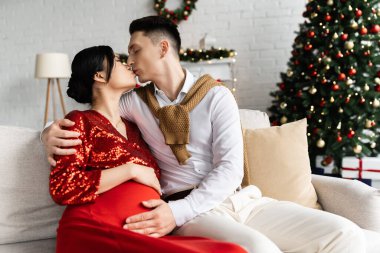 Erkek, hamile Asyalı kadını öpüyor ve karnı burnunda, koltukta, bulanık bir Noel ağacının yanında sarılıyor.