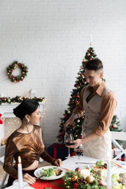 elegant man pouring red wine near asian wife in festive clothes during romantic Christmas supper at home clipart