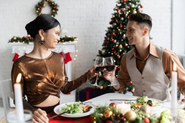 Mutlu ırklar arası çiftler Noel dekoruyla oturma odasında romantik bir akşam yemeğinde kadeh tokuşturuyorlar.