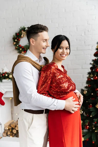 妊娠中のアジアの女性はカメラで笑顔夫は彼女の腹を抱擁しながら彼女の装飾されたリビングルーム — ストック写真